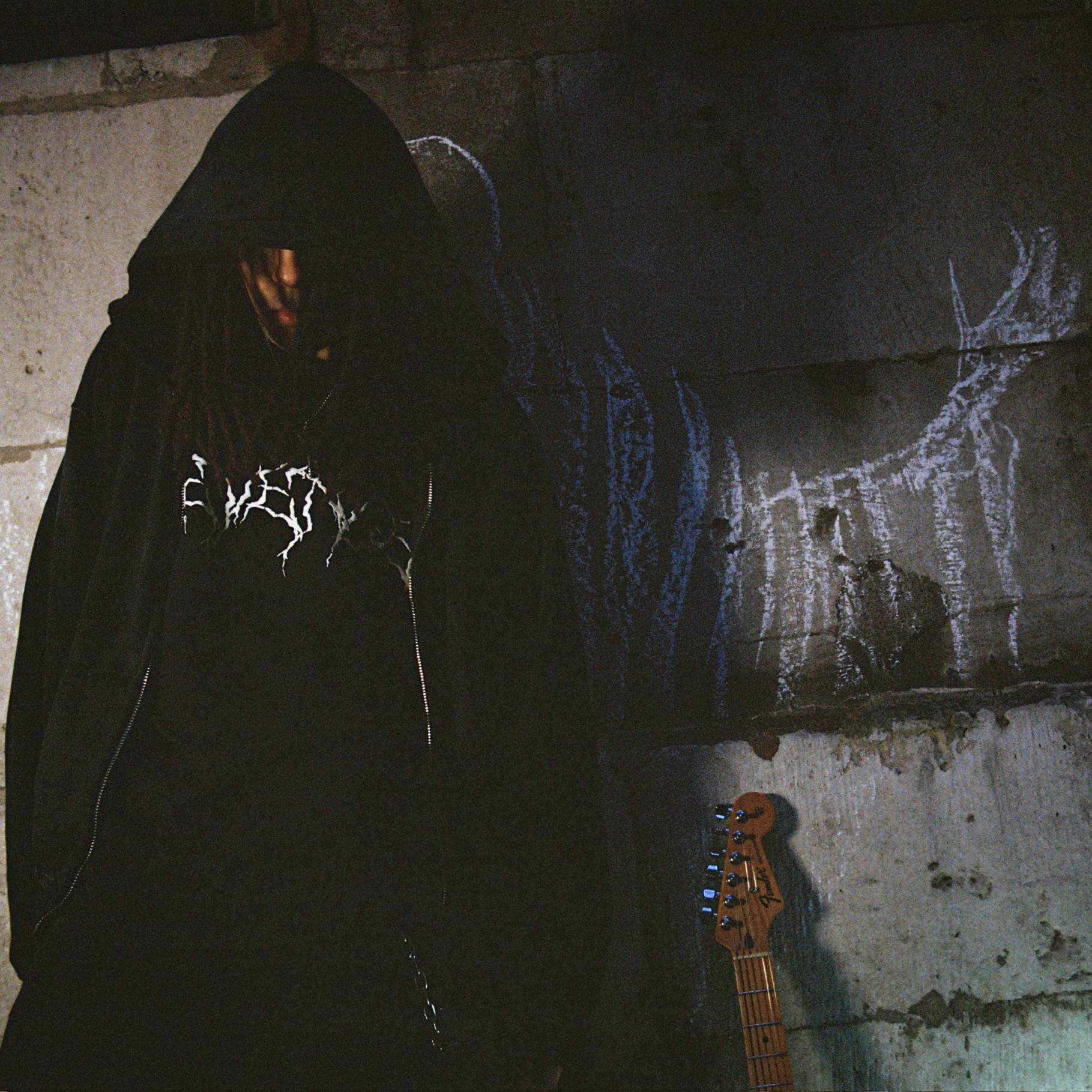 6sezen in a black hood against a chalk-marked concrete wall, a guitar leaning beside him